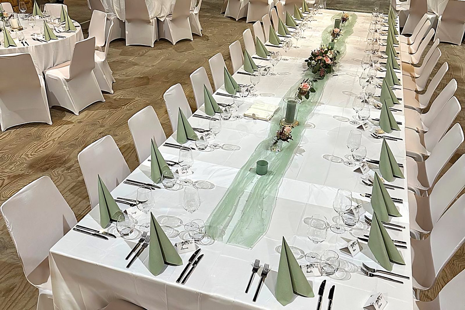 Festtafel mit salbeigrünen Servietten und Blumen in einem Saal mit Holzdachstuhl